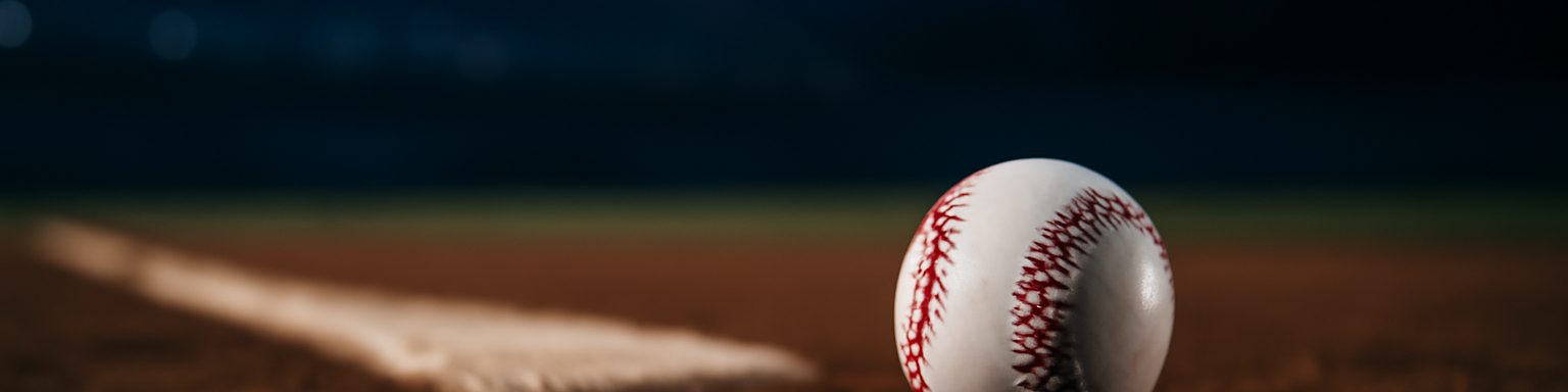 Pelota de béisbol con sus costuras rojas sobre la tierra del cuadro junto a la goma del lanzador, de noche bajo los focos, imagen de esta comparativa de casas de apuestas de béisbol